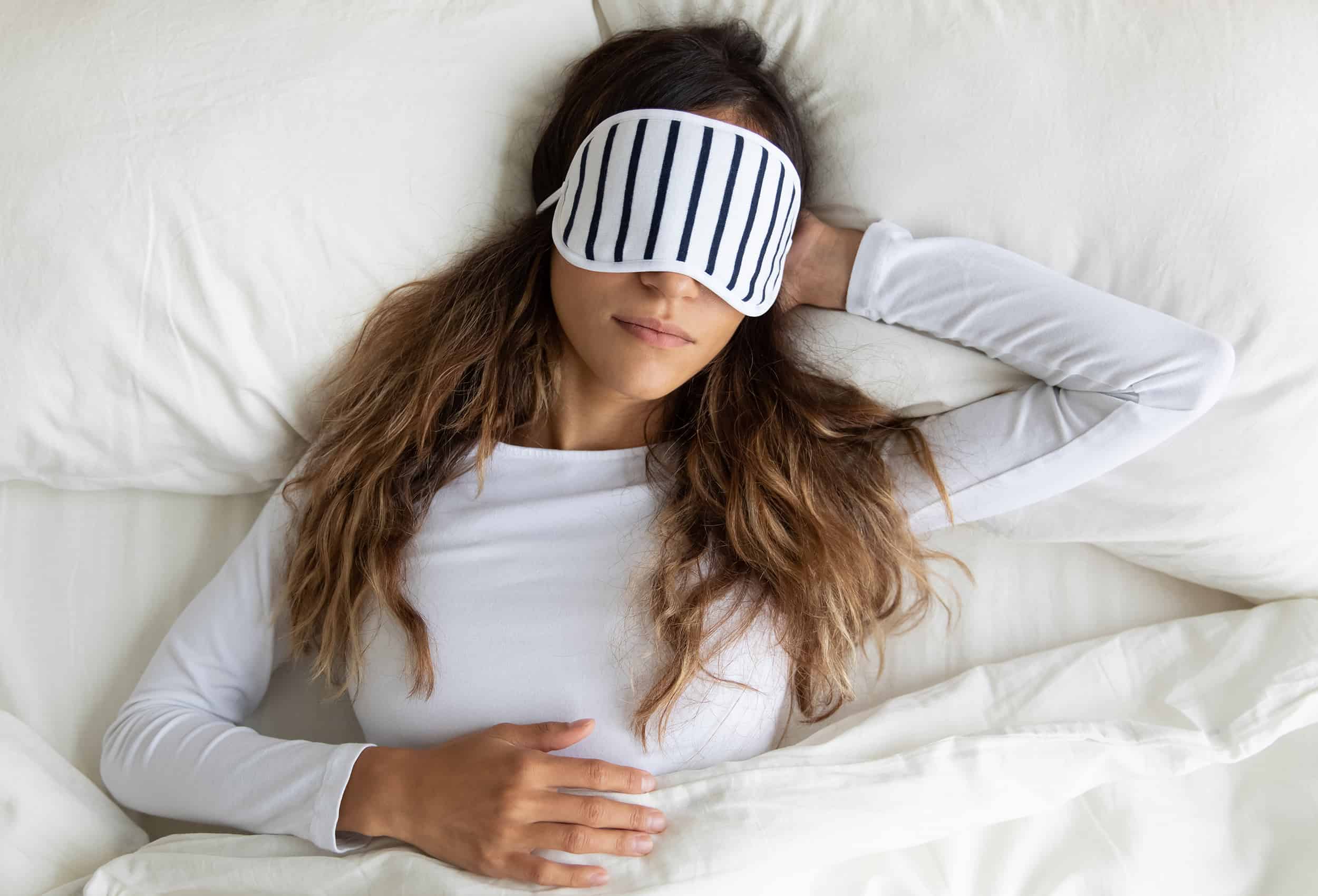 calm-peaceful-woman-wearing-white-and-pin-striped-sleep-mask-laying-in-bed