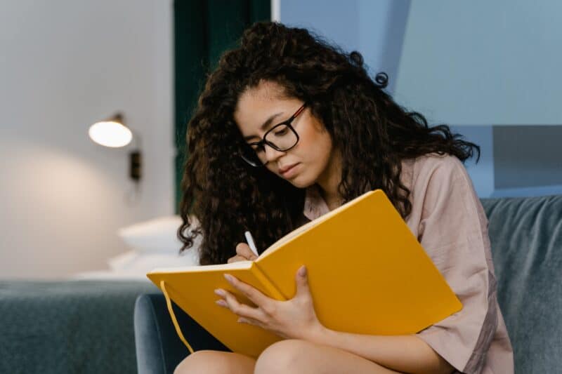 woman-curly-hair-wearing-glasses-writing-in-manifesting-yellow-journal